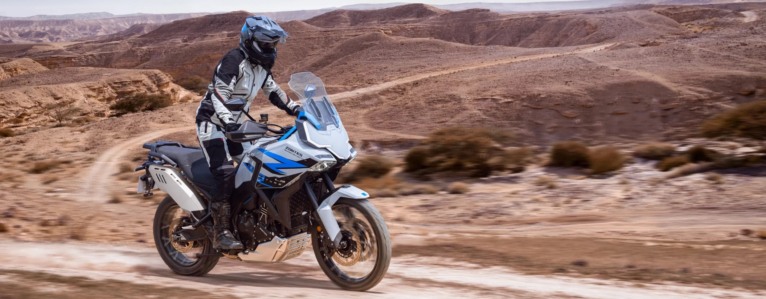 Zontes motos en Argentina - Moto Zontes 703F sobre terreno seco con piloto de aventura y fondo de cerros. Zontes motos en Argentina - Zontes 703F circulando por un camino de tierra en un paisaje árido con conductor equipado.