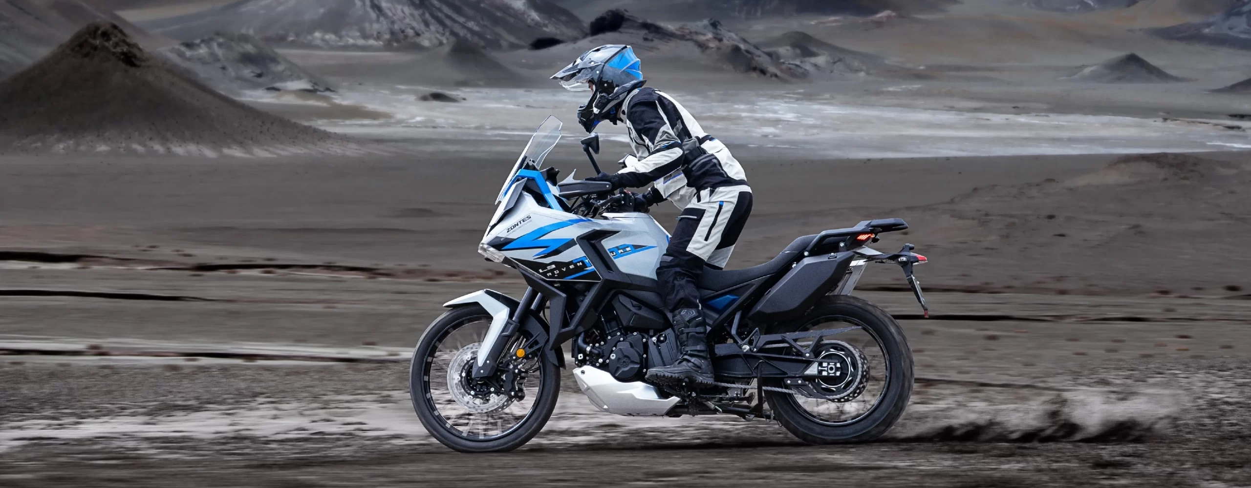Zontes motos en Argentina - Moto Zontes 703F rodando en un terreno abierto con piloto de aventura y paisaje amplio al fondo. Zontes motos en Argentina - Zontes 703F en movimiento sobre suelo oscuro y húmedo con conductor equipado.