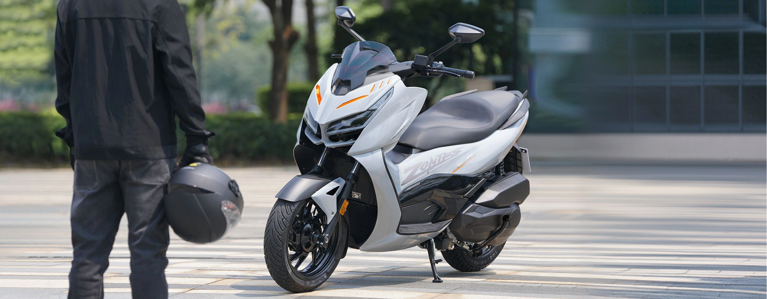 Zontes motos en Argentina - Scooter 368D estacionado con pata lateral desplegada y acompanamiento de conductor equipado. Zontes motos en Argentina - Zontes 368D de carroceria clara detenido sobre una explanada junto a una persona con casco en la mano.